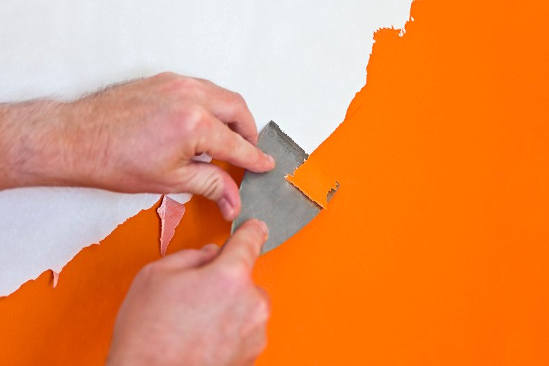 pair of hands removing orange paint off the wall using putty knife